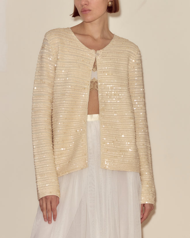 A woman stands against a neutral background wearing the Sequin Spotlight Swing Cardigan in ivory, layered over a lace top and sheer white skirt.