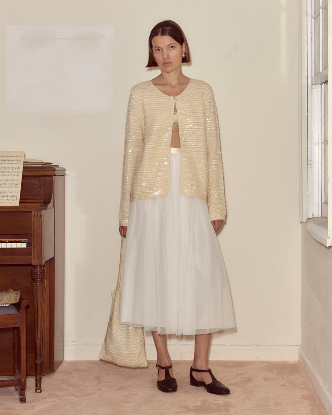 Indoors beside a piano, a woman wears the Sequin Spotlight Swing Cardigan in Ivory, paired with a white tulle skirt and black shoes.