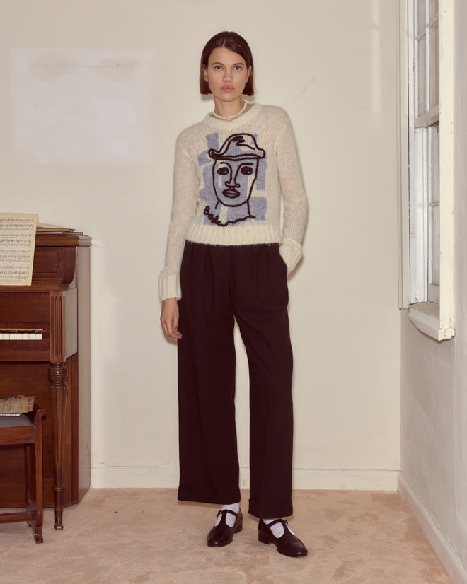 Woman wearing the Chapeau Pullover— a superfine alpaca sweater with an abstract face design, handcrafted in Peru—paired with black pants and shoes, standing by a piano in a bright room.