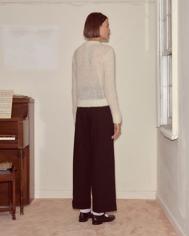 A woman wears the Chapeau Pullover, a cream superfine alpaca knit sweater, paired with black pants as she stands indoors near a piano and a window, facing sideways.