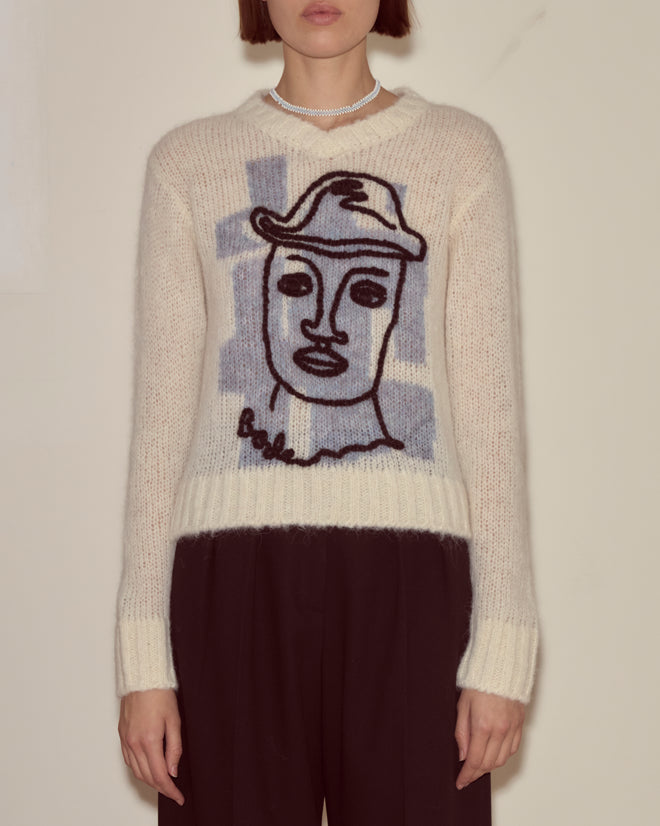 A woman wears the Chapeau Pullover, a cream superfine alpaca sweater featuring a line drawing of a face and hat, made in Peru, paired with black pants.
