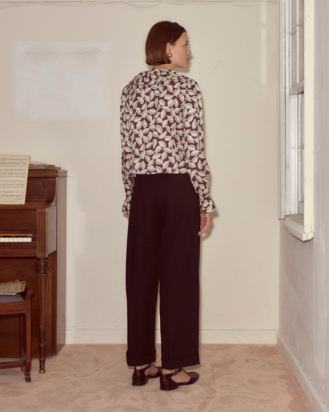 A woman stands indoors by a window and piano, wearing the Paper Birch Flounce Blouse with a leaf-stamp pattern and black pants.