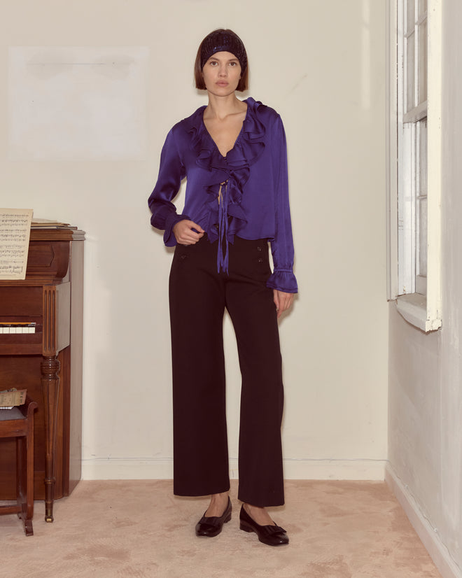 A woman stands indoors by a piano, facing the camera, wearing the Dream Souvenir Flounce Blouse and black pants.