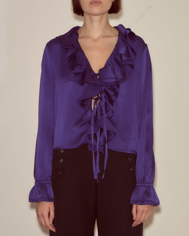 Woman wearing the Dream Souvenir Flounce Blouse, a deep purple silk top with ruffles and a ribbon tie front, paired with black pants against a plain background.