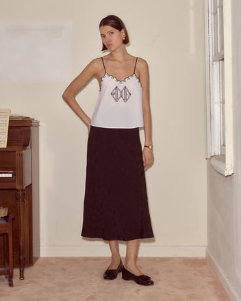 Woman in a Diamond Monogram Tank and black skirt stands indoors near a piano and window, looking at the camera.