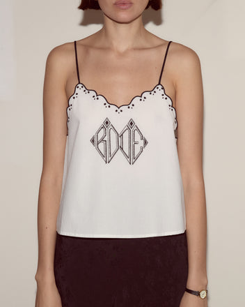 Woman in the Diamond Monogram Tank, a white scalloped embroidered top with black detailing, styled with black pants and a gold watch.