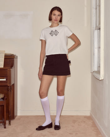 A woman stands in a simple room wearing the Diamond Monogram Tee, a black skirt, white knee-high socks, and black shoes.