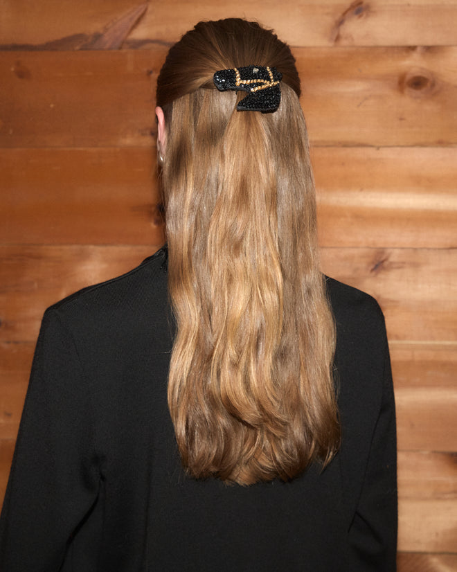 A person with long, wavy, light brown hair wearing the Sequin Pony Barrette in Black stands facing a wooden wall.