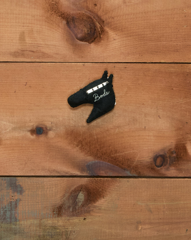 A small black horse head patch, labeled “Sequin Pony Brooch - Brown,” rests on a brown wooden surface with visible knots, evoking the charm of 1940s horse pins.