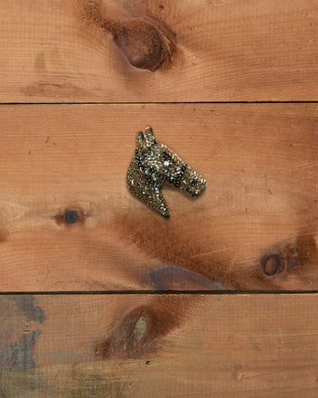 A Sequin Pony Brooch - Brown, featuring a small glittery horse head with a gemstone eye, rests on a wooden surface with visible knots and grain.