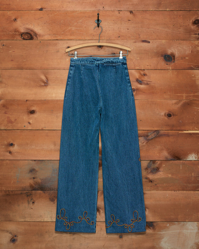 Rawlins Sailor Trousers, featuring a high-waisted blue silhouette with wide legs and brown embroidery on the hem inspired by classic cowboy style, hanging against a wooden wall.