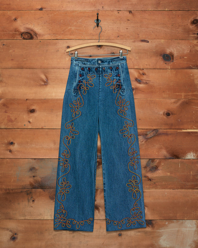Rawlins Sailor Trousers in high-waisted blue denim feature orange floral embroidery and are displayed on a wooden hanger against a wood wall—perfect for pairing with your favorite Western jacket for a complete cowboy-inspired look.