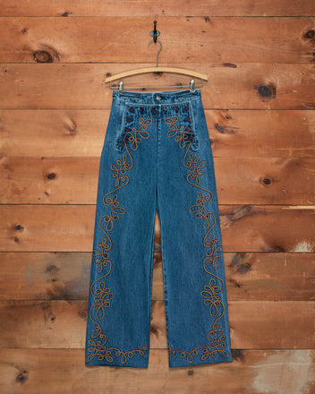 Rawlins Sailor Trousers in high-waisted blue denim feature orange floral embroidery and are displayed on a wooden hanger against a wood wall—perfect for pairing with your favorite Western jacket for a complete cowboy-inspired look.