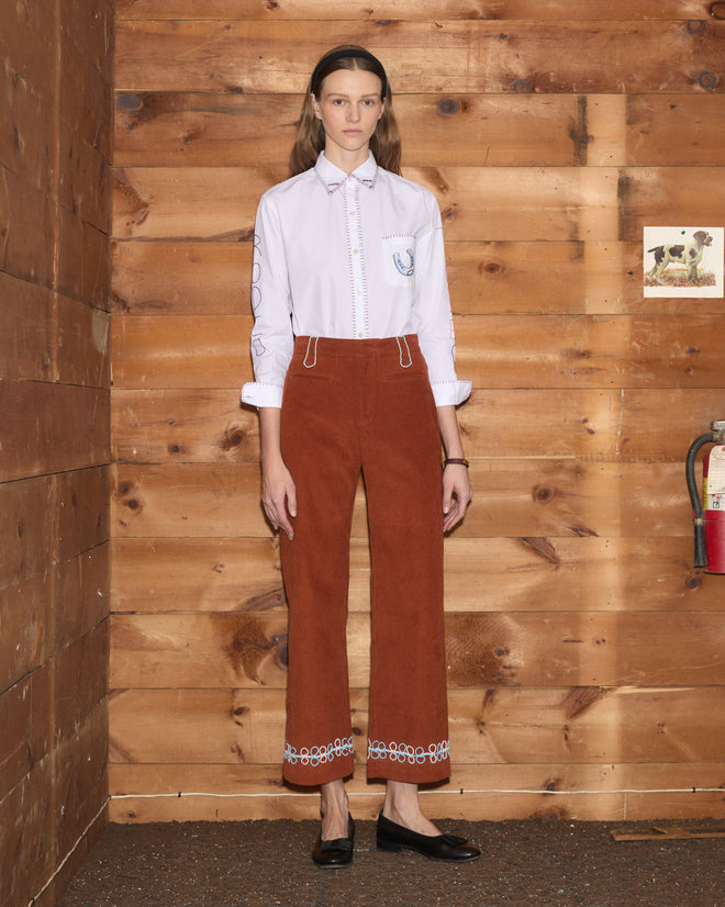 Model wearing a white collared shirt and rust-colored Cheval Corduroy Trousers with a scalloped blue hem and beaded belt loops.