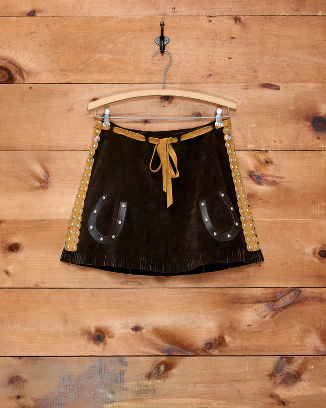 The Carson Skirt—a brown suede mini skirt featuring horseshoe pockets, a fringed hem, and grommet details—hangs on a wooden wall. It’s the perfect choice for a girls’ Western costume.