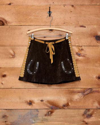 The Carson Skirt—a brown suede mini skirt featuring horseshoe pockets, a fringed hem, and grommet details—hangs on a wooden wall. It’s the perfect choice for a girls’ Western costume.
