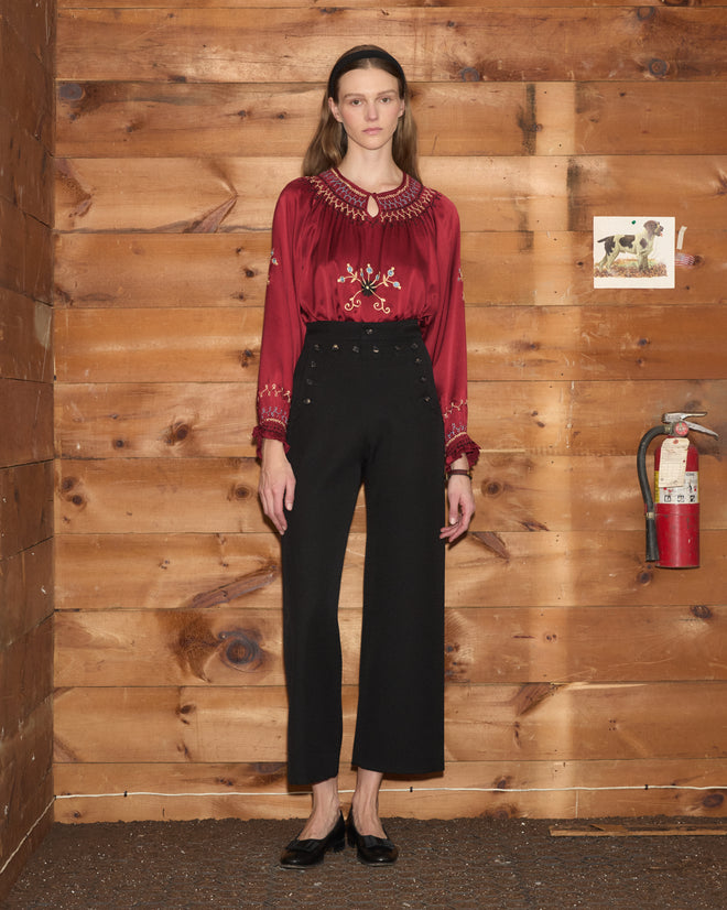 A woman wears a red embroidered blouse and black Riding Twill Sailor Trousers with double-row buttons.
