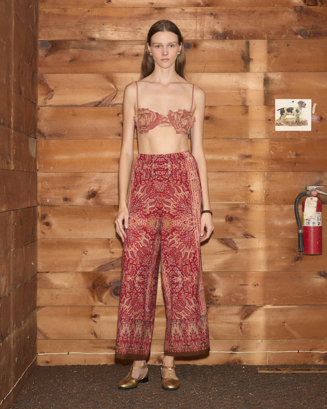 Woman in Arrowleaf Pants and a sheer bra stands against a wooden wall, near a fire extinguisher and photo.