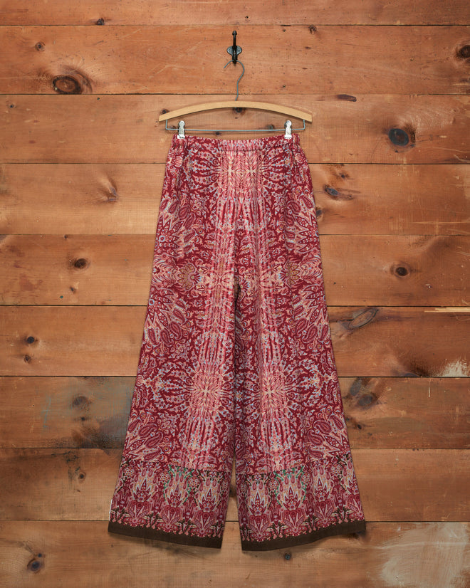 Arrowleaf Pants in a red and pink paisley print, wide-leg style, displayed on a wooden hanger against a wooden wall background.