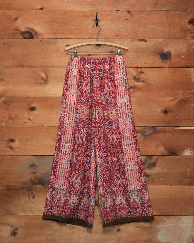 Arrowleaf Pants featuring a red and pink paisley print, displayed hanging on a wooden hanger against a rustic wooden wall.