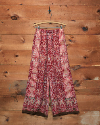 Arrowleaf Pants featuring a red and pink paisley print, displayed hanging on a wooden hanger against a rustic wooden wall.