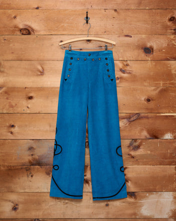 Stagecoach Corduroy Sailor Trousers, crafted from 100% cotton in a blue wide-leg silhouette and finished with a black soutache braid trim, are displayed against a wooden wall background.