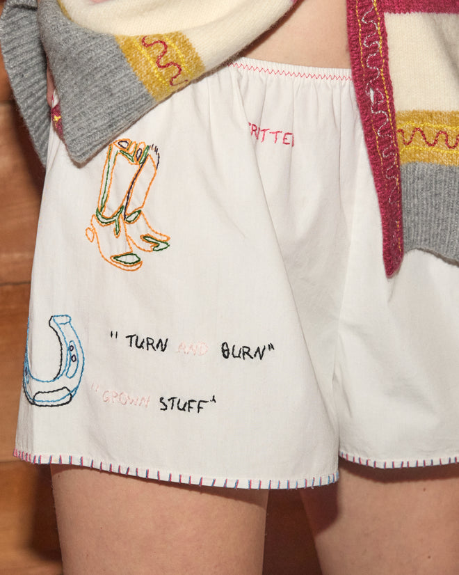 A person wearing the Rodeo Dictionary Tap Shorts—white cotton poplin shorts featuring colorful embroidered designs and phrases, styled as women’s western shorts—paired with a patterned sweater.