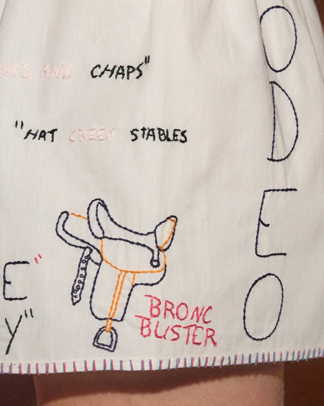 Introducing the Rodeo Dictionary Tap Shorts: crafted from white cotton poplin and featuring hand-embroidered text, a detailed saddle motif, and the word “RODEO” boldly displayed in large blue letters along the side. These stylish shorts are the perfect addition to any collection of women’s western shorts.
