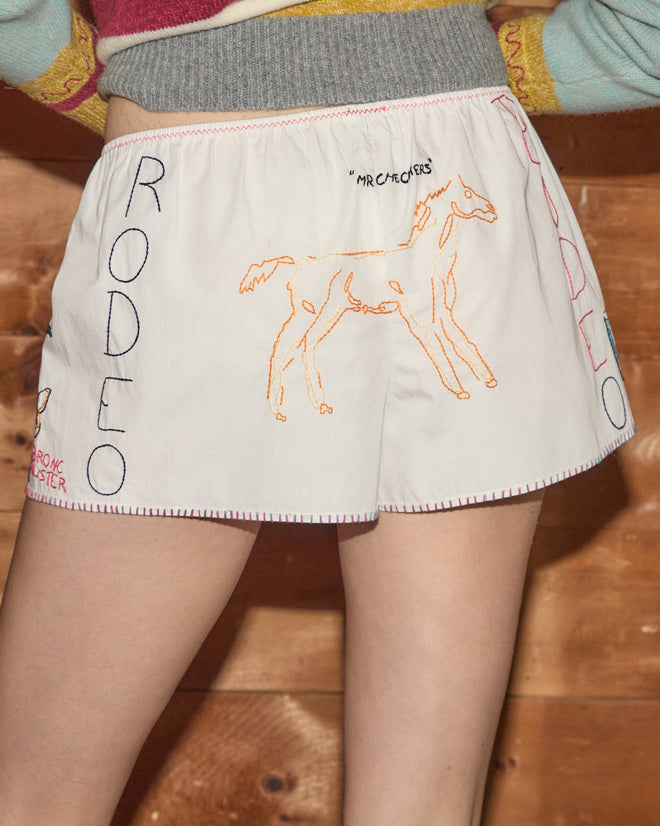 Wearing the Rodeo Dictionary Tap Shorts—white women’s western shorts made from soft cotton poplin and hand-embroidered with a “RODEO” and horse motif—the person stands in front of wood paneling, showing off comfort and charm.