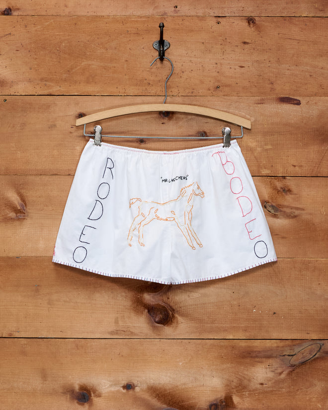 Rodeo Dictionary Tap Shorts—white boxer-style shorts with hand-embroidered horse and “Rodeo” text, displayed against a wooden wall, blend western charm with effortless style.
