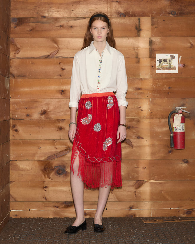 A woman in a white shirt and the Sundance Skirt stands indoors by a wood wall, next to a fire extinguisher and a dog photo.