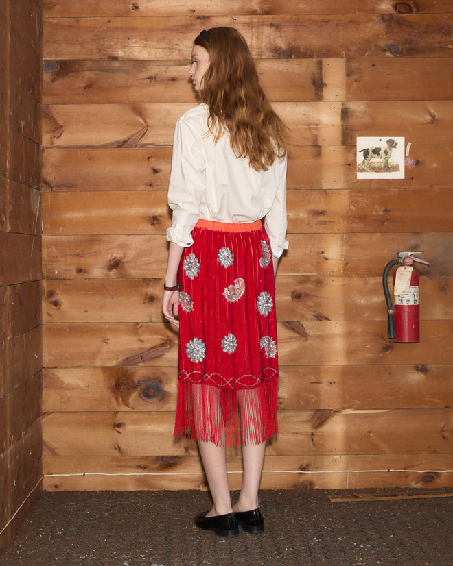 A woman with long hair wearing a white blouse and the Sundance Skirt—a scarlet sequin skirt with an asymmetrical hem—stands facing a wooden wall with a fire extinguisher.