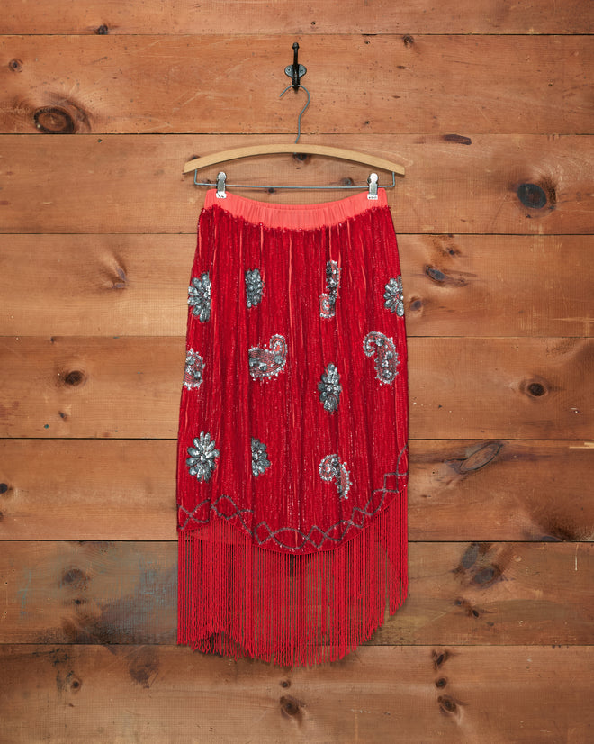 The Sundance Skirt features scarlet sequins with shimmering silver designs and an asymmetrical fringe hem, shown here hanging on a wooden hanger against a rustic wood wall—ideal for creating a striking burlesque costume look.