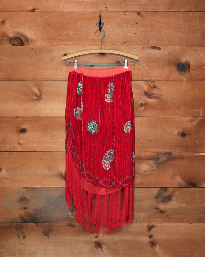 Sundance Skirt—a scarlet sequin skirt featuring shimmering silver fringe designs and an asymmetrical hem, displayed on a wooden hanger against a wooden wall—makes a striking choice for a burlesque costume.