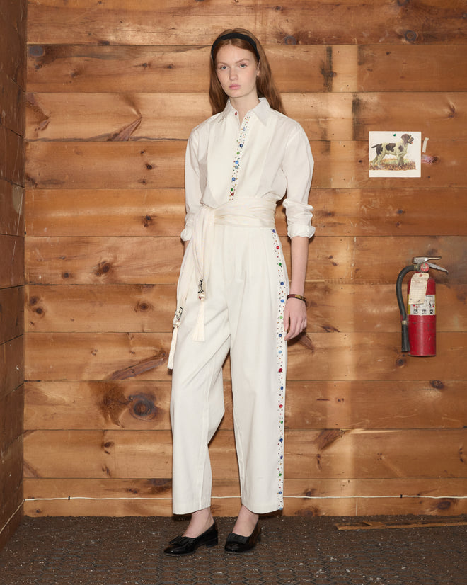 A woman in a white shirt and Button Jar Trousers stands by a wooden wall with a fire extinguisher and a small dog photo.