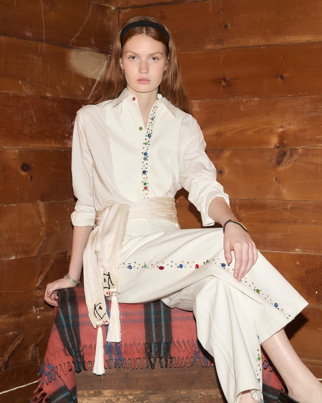 A woman wearing the Button Jar Trousers sits on a blanket-covered bench against a wooden wall.