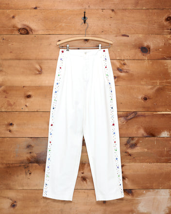Button Jar Trousers—white high-waisted cotton trousers featuring vibrant floral embroidery along the sides—hang on a hook against a wooden wall.