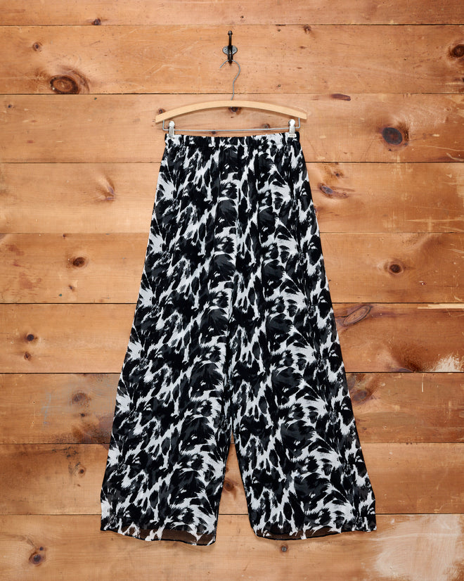 The Appaloosa Silk Pants—wide-leg and crafted from luxurious silk crepe, featuring a bold black and white abstract pattern inspired by Appaloosa spots—hang against a wooden wall.