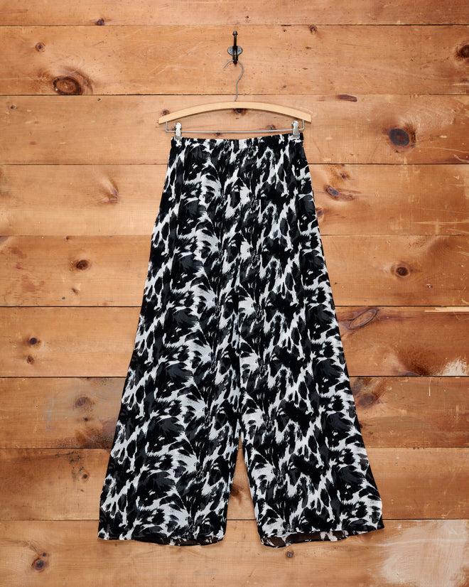 Appaloosa Silk Pants featuring a black and white spots pattern and wide-leg silhouette in silk crepe are displayed on a hanger against a wooden wall background.