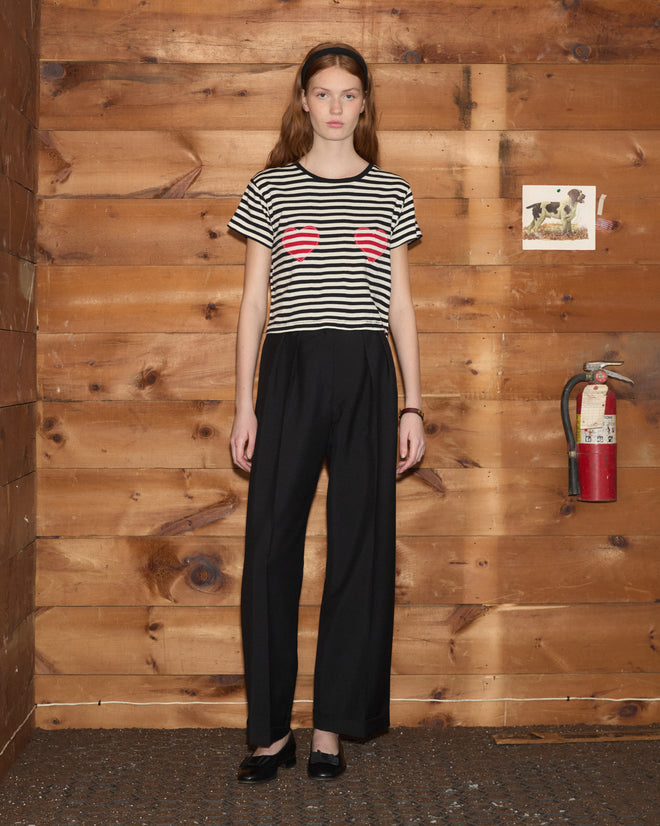A woman wearing the Rib Stripe Heart Tee and black pants stands indoors near a wood wall and a fire extinguisher.