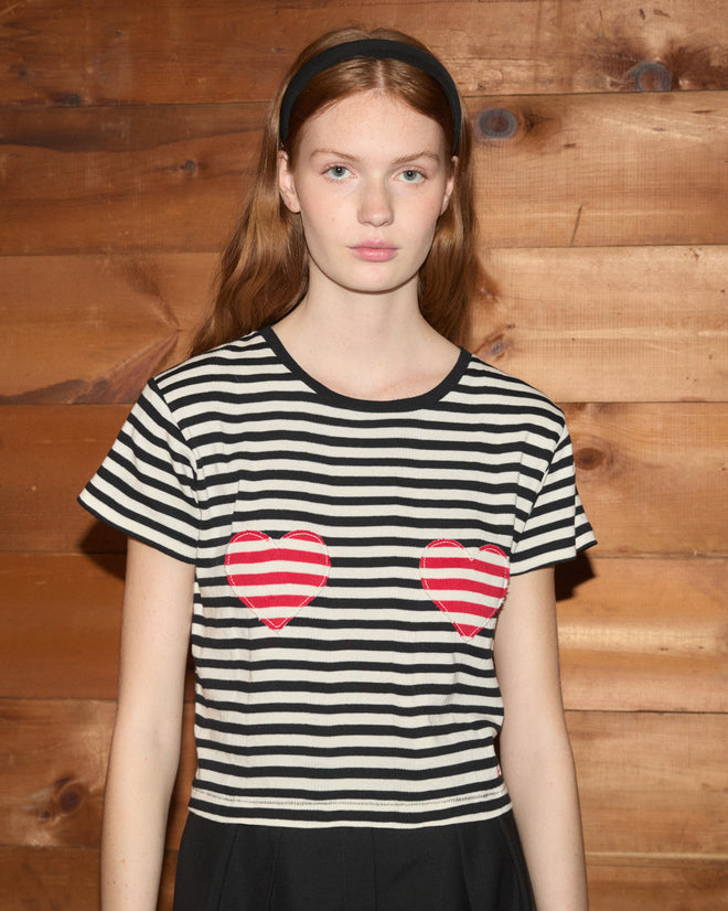 A woman wearing the Rib Stripe Heart Tee, crafted from 100% cotton and adorned with red hearts, stands against a wooden wall, looking at the camera.