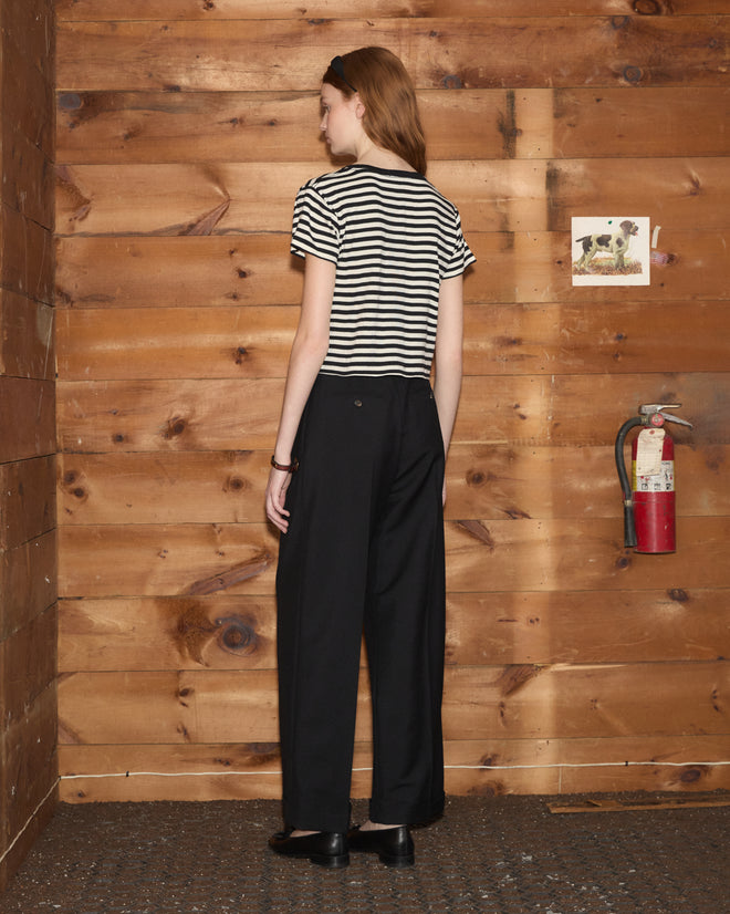 A woman wearing the Rib Stripe Heart Tee and black pants stands facing a wooden wall with a fire extinguisher and photo.