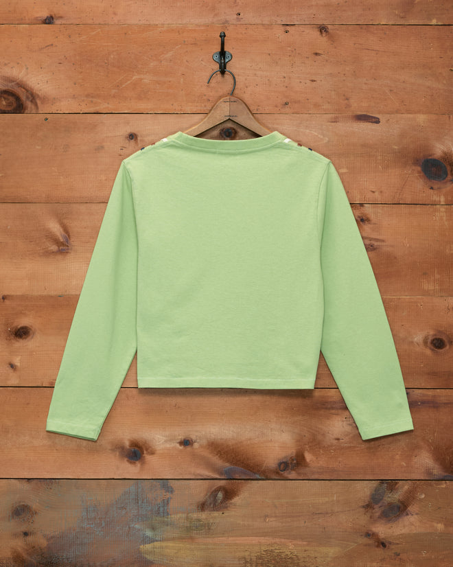 The Sidewinder Long Sleeve Tee in light green, made from 100% cotton, hangs on a hanger and is displayed from the back against a wooden wall background.