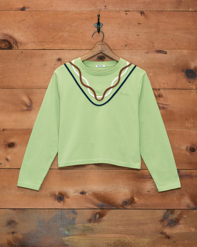 Sidewinder Long Sleeve Tee in light green, crafted from 100% cotton and featuring a wavy, multicolored neckline inspired by classic 1960s sportswear, displayed on a wooden wall.
