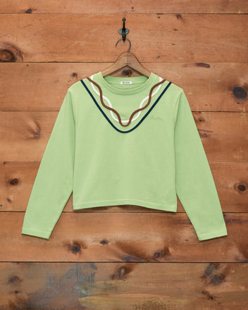 Sidewinder Long Sleeve Tee in light green, crafted from 100% cotton and featuring a wavy, multicolored neckline inspired by classic 1960s sportswear, displayed on a wooden wall.