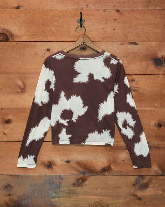Featherweight Cow Print Long Sleeve Tee in brown and white, featuring a vintage-inspired cow pattern, lightweight fabric, cropped fit, and long sleeves—displayed on a wooden wall.
