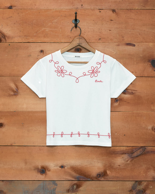 Lasso Tee, a white 100% cotton t-shirt with pink floral and loop couching embroidery, made in Portugal, displayed on a wooden hanger against a wood wall.