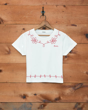 Lasso Tee, a white 100% cotton t-shirt with pink floral and loop couching embroidery, made in Portugal, displayed on a wooden hanger against a wood wall.