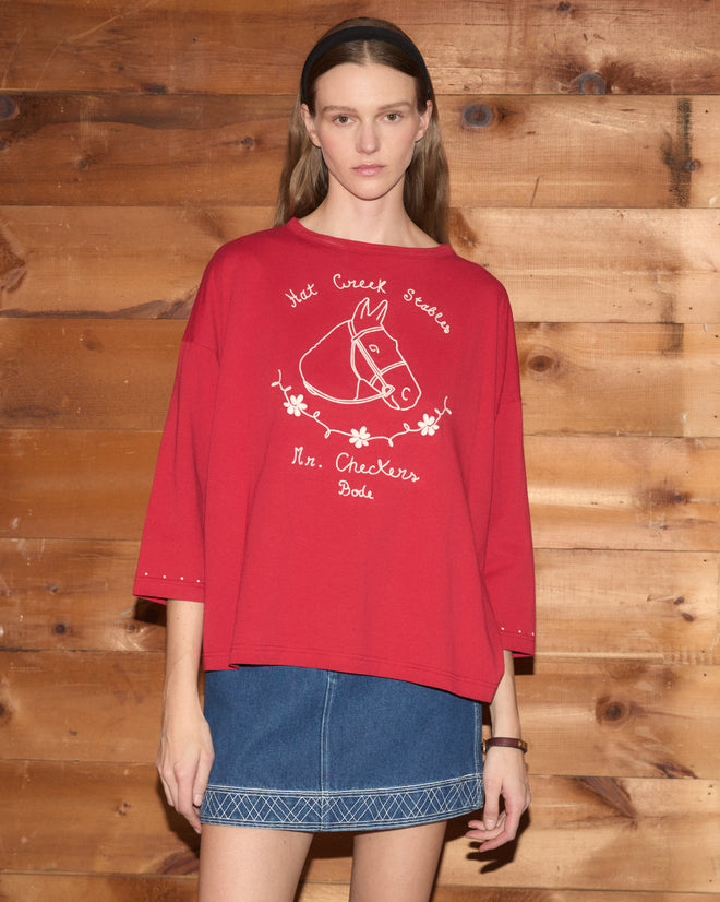 A woman stands in front of a wooden wall, wearing the Stables Long Sleeve Tee—a red raglan shirt with a horse graphic, crafted from 100% cotton—paired with a denim skirt.