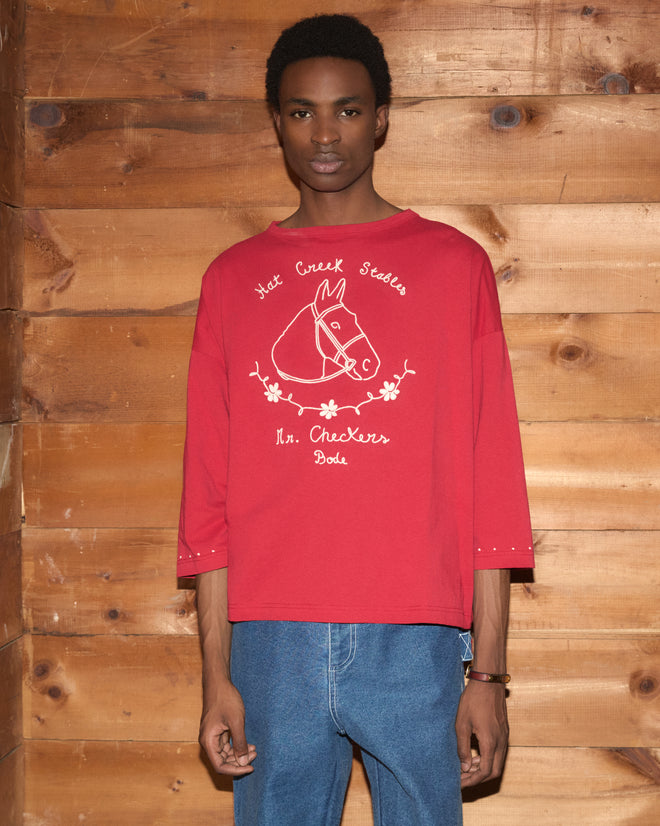Wearing the Stables Long Sleeve Tee—a 100% cotton red raglan shirt with a horse graphic and detailed chain-stitching—the person stands in front of a wooden wall.